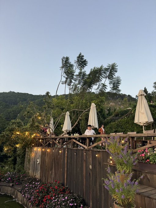 An outdoor wooden deck or patio area with closed umbrellas, surrounded by trees and flowers, where two people can be seen in the background during what appears to be dusk.