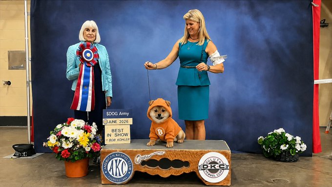 The image features a woman holding an orange dog, possibly dressed up for a competition or event. They are standing next to a table with a sign that reads "$DOG is Best in Show (June 2026)". The background of the photo shows a blue wall and potted plants, creating a pleasant atmosphere around them.