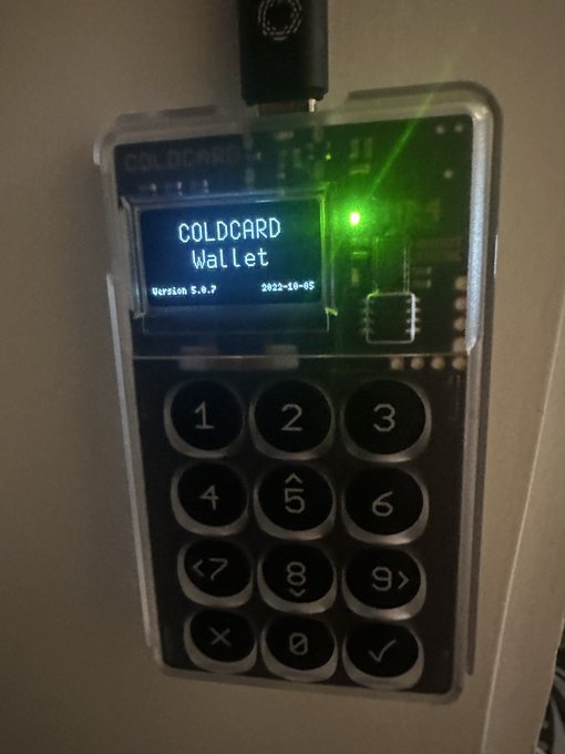 A powered-on COLDCARD Wallet displaying 'COLDCARD Wallet, Version 5.0.7, 2022-10-05' with a glowing green LED, plugged in via USB.