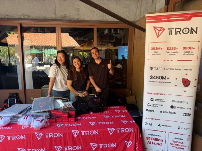 Three TRON representatives pose behind a branded table at Stanford's Science of Blockchain Conference, with the banner highlighting key stats like "393M+ User accounts," "$23B+ Daily transfer volume," "$90B+ USDT in circulation," and a "$1 Billion AI Fund" application. The setup, complete with TRON-branded merchandise and partnerships like T3FCU, Tether, and Mastercard, visually reinforces the post's message about TRON's active engagement with students and builders in the blockchain space.