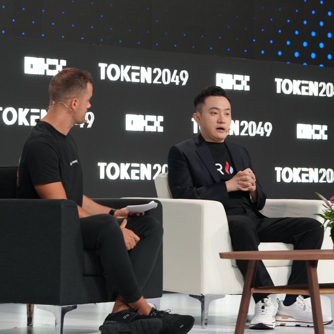 Justin Sun speaks during a TOKEN2049 panel discussion, with the event's logo displayed prominently behind him and the interviewer, discussing how younger generations are moving away from traditional banking.