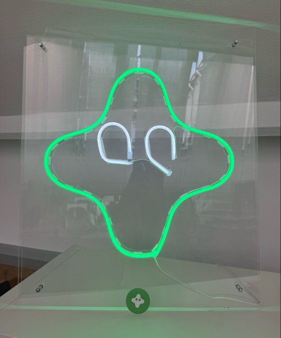 A green neon sign shaped like a star with the abstract outline of a face, displayed on a clear acrylic panel with a small green circular button at the bottom. The post "gUmia" appears to be a stylistic reference, matching the design's aesthetic branding.