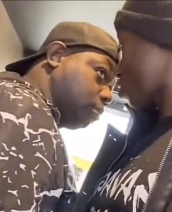 Two men in dark clothing stand face-to-face in a close, intense moment, with one wearing a cap and patterned shirt.