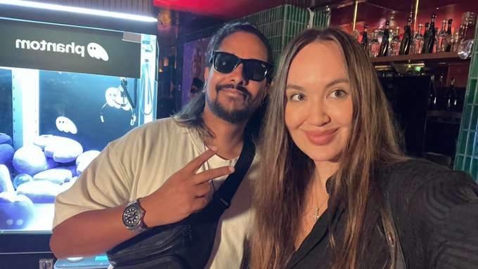A man with long hair and sunglasses and a woman with long brown hair take a selfie in front of a claw machine with a reversed logo reading 'priSnigm' and a bar in the background.