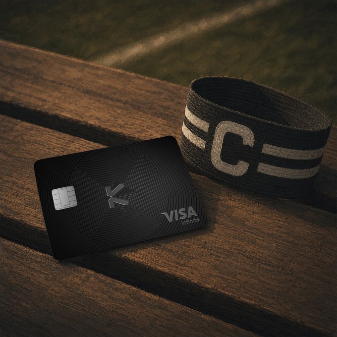 A black Visa Infinite metal credit card with a 'K' logo rests on a wooden slatted surface next to a black captain's armband with a white 'C' insignia.