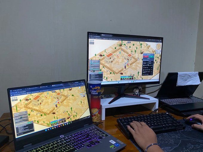 A person is grinding the Web3 game "Kintara" (@PlayKintara) on a dual-monitor setup with a laptop and an external display, both showing the same isometric desert village game. Their hands rest on the keyboard and mouse alongside a can of "Chips Oman" snacks, capturing a casual co-op gaming session while waiting for the "King Scorpo" to drop.