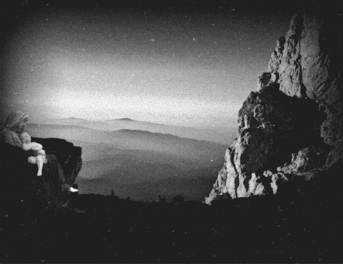 A vintage black-and-white photograph showing a silhouetted figure with a child sitting on a rocky cliff, gazing out over a misty mountain range at twilight with faint stars visible in the sky.