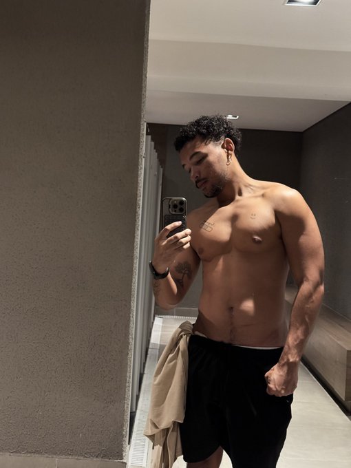 A shirtless man takes a mirror selfie in what appears to be a bathroom, showing a toned physique, dark shorts, and a towel draped over his shoulder.