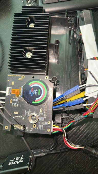 A photograph of computer hardware, featuring a circular OLED display showing a colorful gauge, a black finned heatsink, and various connected cables including a SATA power connector.