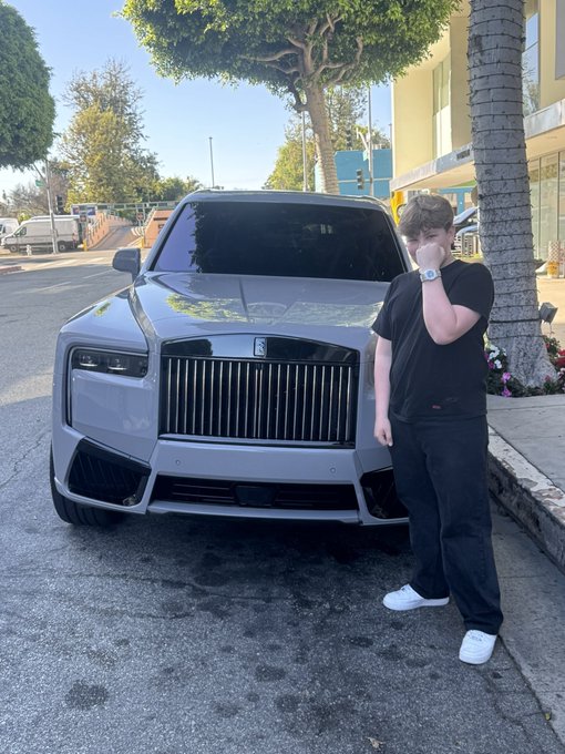 A young person in a black t-shirt, dark pants, and white sneakers stands beside a silver Rolls-Royce Cullinan SUV parked on a street, touching their face.