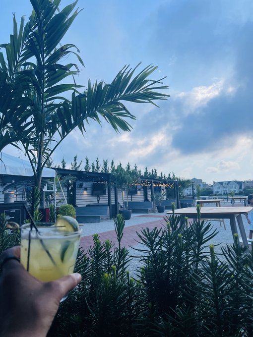 A hand holds a glass of lemonade with mint and lime in the foreground, with a beachside restaurant, palm trees, and cloudy sky in the background.