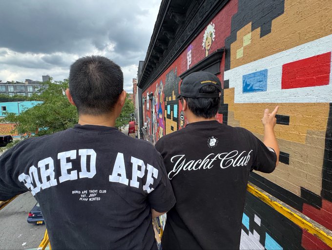 Two people from behind wearing black t-shirts, one reading 'Bored Ape Yacht Club' and the other 'Yacht Club', standing in front of a colorful pixel-art mural.