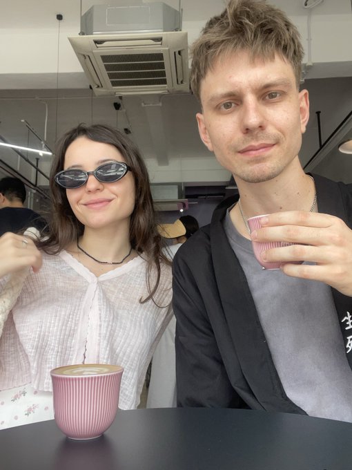 A selfie of a young couple at a cafe, with the woman wearing sunglasses and a white shirt, and the man holding a pink drink, with a pink ribbed cup on the table.