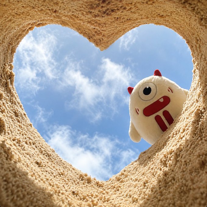 A plush white monster character with red horns peeks out from a sand-colored heart-shaped opening against a backdrop of blue sky and wispy clouds. The post "#TRON" likely refers to the cryptocurrency project TRON (TRX), whose mascot is a similar adorable white character, fitting the cheerful "GM" (Good Morning) greeting.