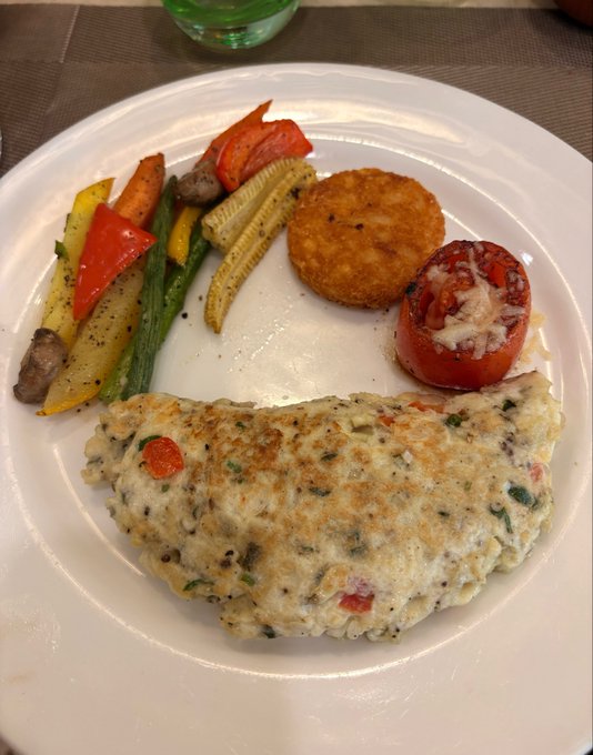 A plated meal featuring a creamy herb-flecked chicken cutlet, roasted vegetables (baby corn, bell peppers, mushrooms), a stuffed tomato, and a crispy potato cake sits on a white plate, paired with a "gm" post that hints at the blogger's love for delicious food and self-care.