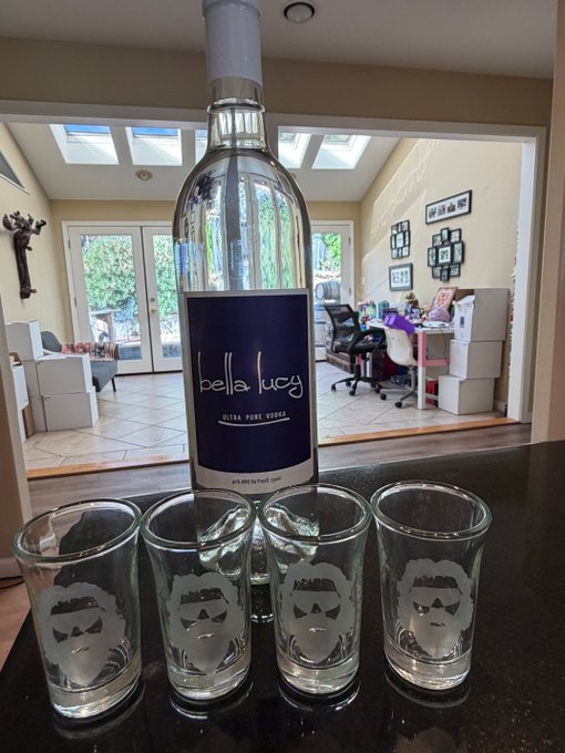 A bottle of "Bella Lucy Ultra Pure Vodka" displayed on a kitchen counter alongside four shot glasses etched with a bearded man's face design, likely the "Mullet Man" NFT collection referenced in the post. The setup showcases the collaboration between the vodka brand and the artist, ready for an upcoming promotional drop.
