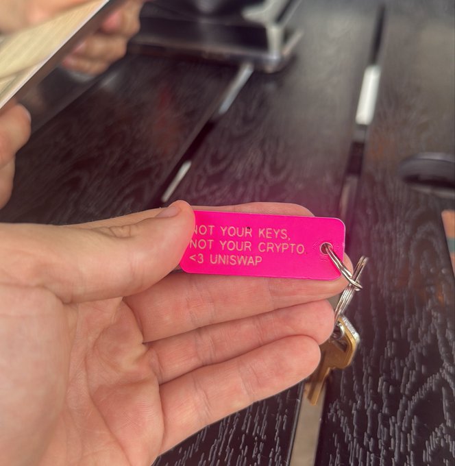A hand holds a bright pink keychain engraved with "NOT YOUR KEYS, NOT YOUR CRYPTO <3 UNISWAP," attached to a set of keys—visually reinforcing the famous crypto self-custody mantra while showing brand loyalty to Uniswap.