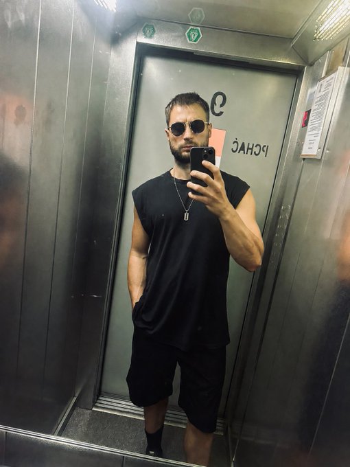 A man in a black sleeveless shirt, black shorts, and sunglasses takes a mirror selfie with his phone inside an elevator with a 'CHAO' sign on the door.