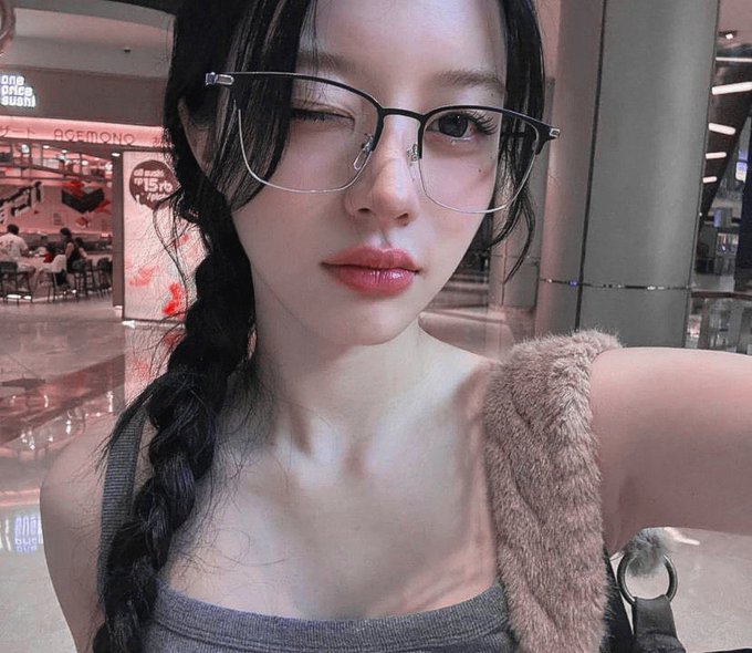 A woman with glasses, a long dark braid, and pink lipstick winks at the camera in a selfie taken inside a shopping mall.