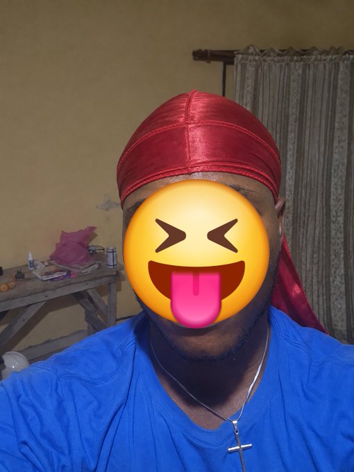 A person wearing a red satin durag, blue shirt, and cross necklace has their face covered with a playful emoji sticker sticking its tongue out, in what appears to be a room with yellow walls.