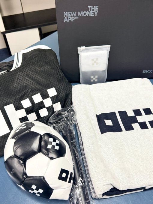A merchandise bundle from OKX labeled 'THE NEW MONEY APP™', including a black and white checkered backpack, a white towel, a small bag, and a black and white OKX branded soccer ball.