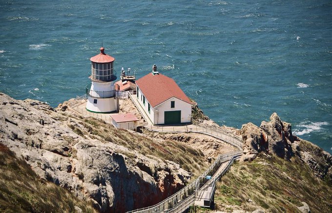 A lighthouse with a red roof and an adjacent building sit on a rocky cliff overlooking the ocean, with a winding staircase leading down the cliffside.