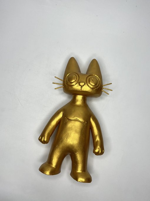A small, hand-painted gold figurine of a stylized cat with large round eyes, whiskers, and pointed ears, photographed against a plain white background.
