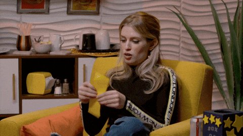 The image shows a woman sitting in an armchair, holding a yellow object or ribbon in her hands while looking at something off-camera. She appears to be engaged in a conversation with someone who is not visible in the frame. The background features various objects such as a potted plant and multiple cups on shelves, suggesting that she might be in a living room setting.
