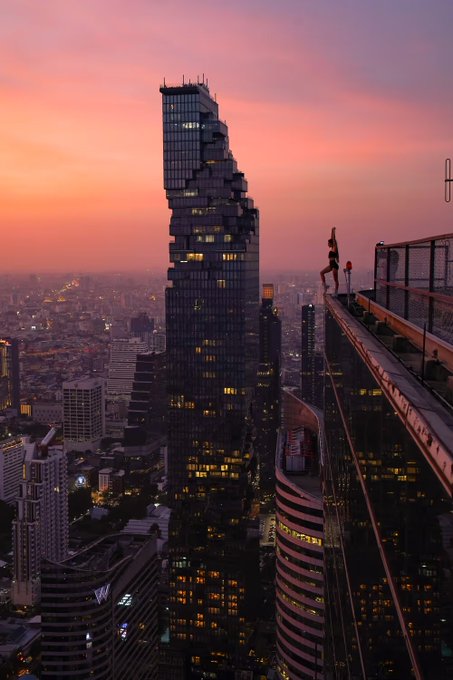 A dramatic cityscape photograph at sunset showing a person standing on the edge of a rooftop, with a tall distinctive Mahanakhon tower in the background against a pink and orange sky.