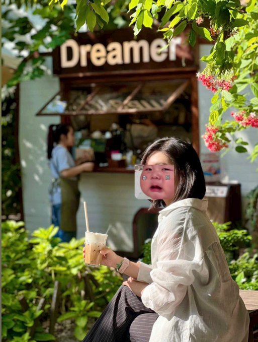 A young woman enjoys a refreshing drink while sitting near a wooden kiosk labeled "Dreamers," surrounded by lush greenery and pink flowers, with a vendor working in the background. The image evokes the post's question about simple, peaceful moments—like savoring a quiet afternoon café visit—that one would pursue after achieving financial success.
