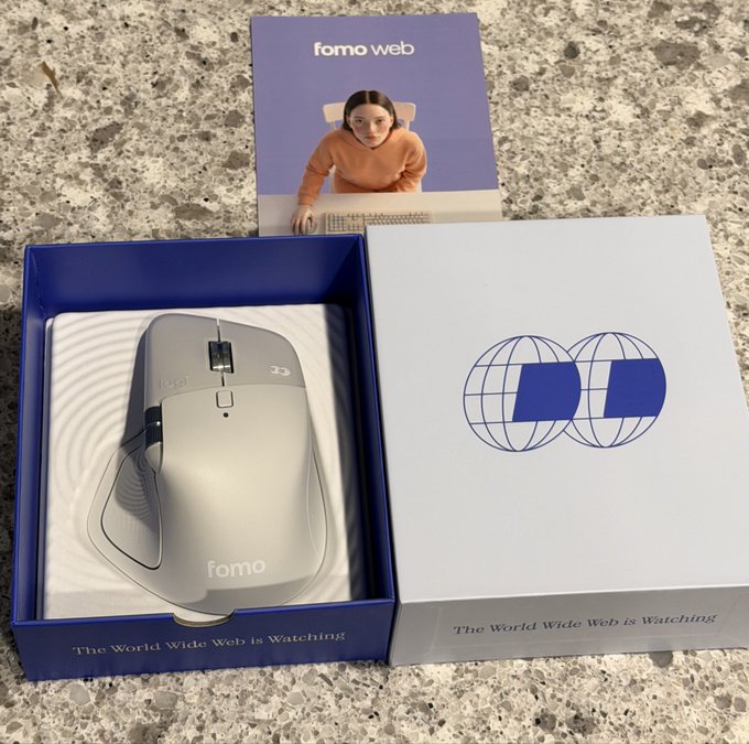 The image features a silver mouse with a blue box containing an open laptop computer, along with a book titled "Famo Web" that has a picture of a woman sitting at a desk. The text on the book reads "No team does it quite like @fomo". This suggests that the image is related to the concept of using technology for global domination or spreading awareness about a particular cause through social media, as indicated by the title of the book and the presence of the laptop in the box.