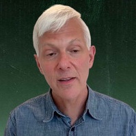 A close-up portrait of an older man with white hair wearing a blue button-up shirt against a dark green background.