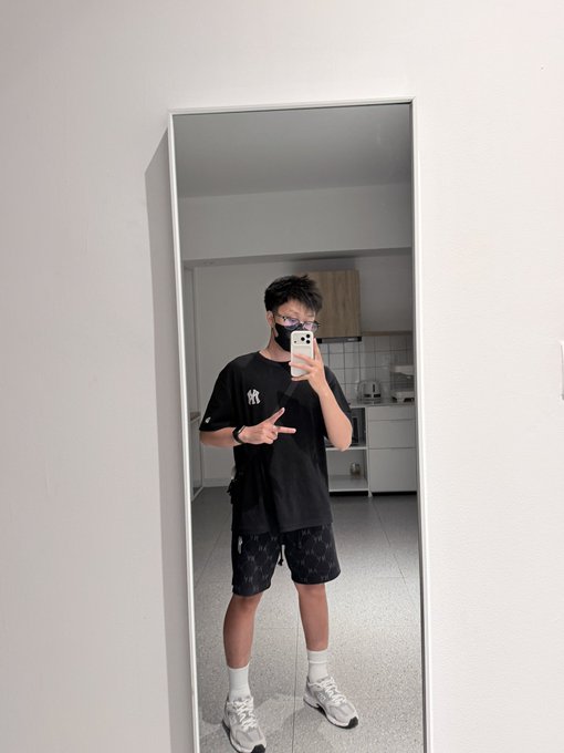 A young man wearing glasses and a mask takes a mirror selfie, making a hand gesture while wearing a black t-shirt and shorts.