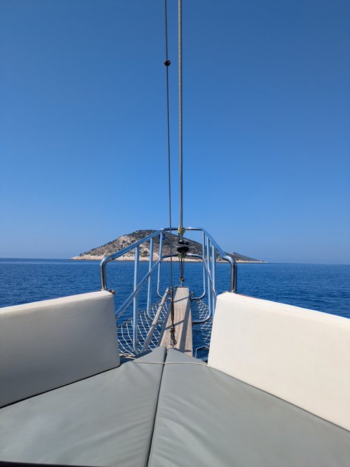 A view from the bow of a sailboat looking out over blue ocean water toward a distant hilly island under a clear sky.