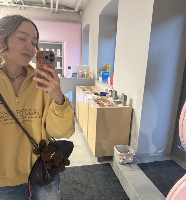 A woman takes a mirror selfie with a wooden phone case, wearing a yellow zip-up hoodie and a small brown teddy bear crossbody bag inside a brightly colored store.