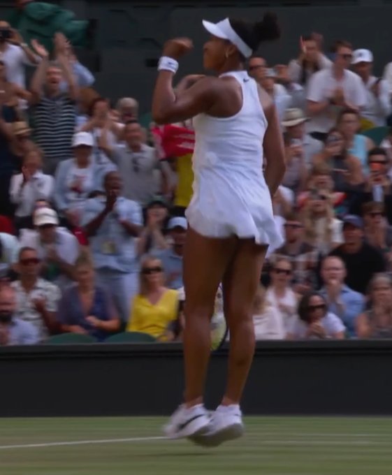 A female tennis player in a white outfit jumps in celebration on a grass court, pumping her fist in triumph as the crowd cheers behind her, suggesting a hard-fought victory at Wimbledon.