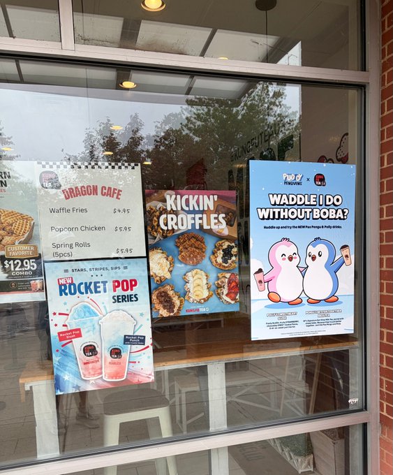 A storefront window displaying posters for Dragon Cafe menu items like Waffle Fries and Kickin' Croffles, alongside promotional flyers for a new Rocket Pop drink series and a 'Waddle I Do Without BOBA?' Pudgy Penguins collaboration.