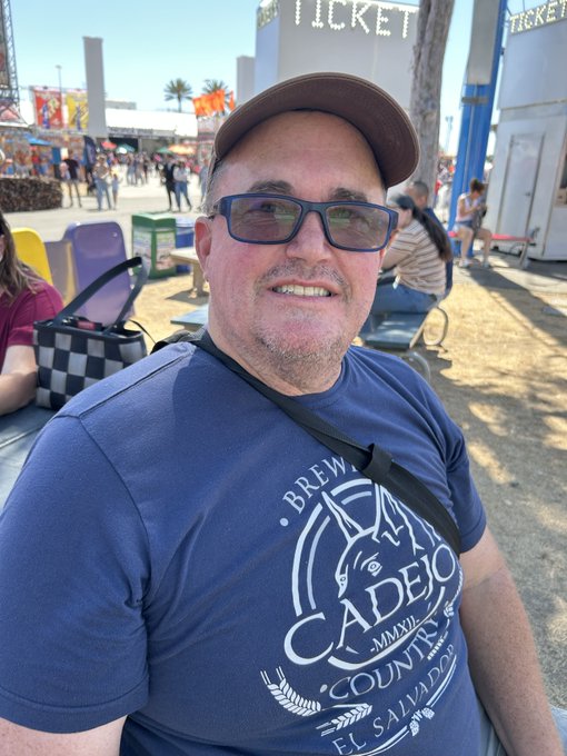 A man wearing a cap, sunglasses, and a blue 'CADEJO COUNTRY' t-shirt smiles for a selfie at an outdoor festival with ticket booths and attendees in the background.