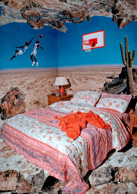 The image features an orange bedspread with white flowers that covers a bed, along with two pillows. The bed is situated next to a window overlooking a desert landscape. A basketball hoop can be seen in the background, suggesting that this room might be part of a sports-themed hotel or motel. The post also mentions GM and happy Monday, which could indicate that the image was taken during the weekend or a holiday when people are more likely to travel for leisure purposes.
