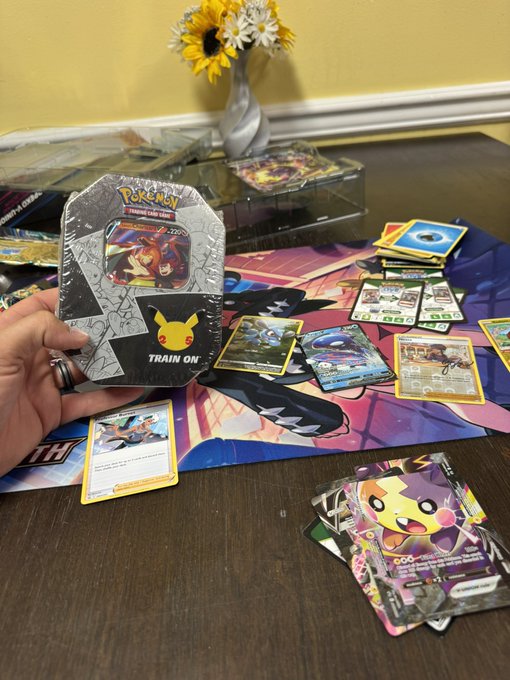 A person holding a sealed Pokémon Trading Card Game tin ('TRAIN ON') over a colorful playmat scattered with various Pokémon cards and booster packs, with sunflowers in a vase in the background.