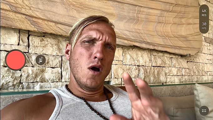The image shows a man wearing a white tank top, sitting next to a couch with his hand raised as if he's about to make a point or ask a question. The background features a brick wall that adds an interesting visual element to the scene. The caption of the image reads "bitcoin: we need to talk. NOW. youtu.be/y5AZoPQYrUg [click]". This suggests that there might be some sort of discussion or conversation taking place, possibly related to Bitcoin or a similar topic.