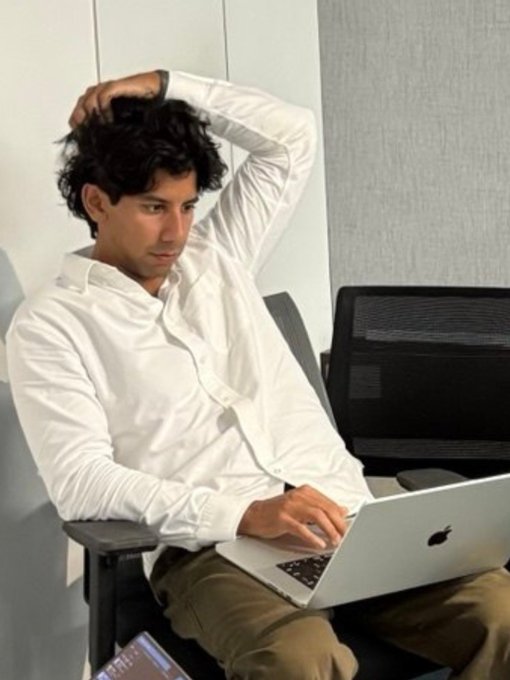 A young man with curly dark hair wearing a white button-up shirt, sitting in a chair while using a MacBook laptop with one hand behind his head.
