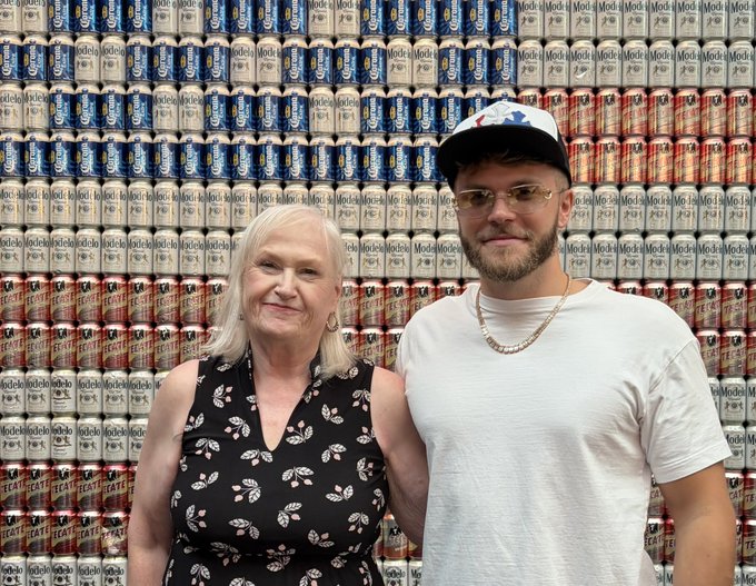 A woman in a black butterfly-patterned dress standing next to a man in a white t-shirt, sunglasses, and a baseball cap, posing in front of a wall of stacked beer cans (Modelo and Tecate).