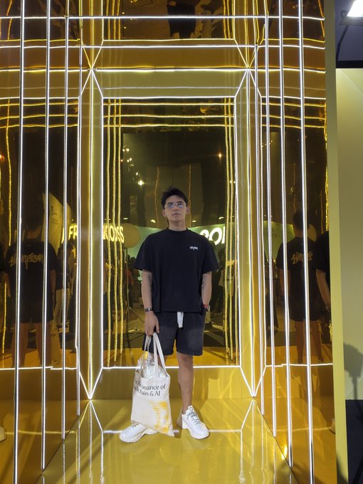 A young man wearing glasses, a black t-shirt, and shorts poses in a mirrored hallway illuminated by yellow and white LED light strips, holding a shopping bag labeled 'Assurance of NICALOI'.
