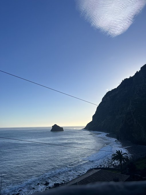 A serene coastal view at dawn shows a rocky island silhouetted against the calm ocean, with steep cliffs and a wire cutting across the foreground, while the message "Gm familia say it back" greets followers with a peaceful morning greeting matching the tranquil seaside scene.