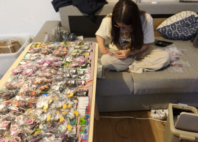 A woman sits cross-legged on a couch carefully assembling colorful acrylic charms, with a large table covered in bagged character keychains and craft supplies in front of her. The image illustrates the hands-on production work behind Silly Kitties, highlighting the co-founder's dedication to handcrafting the company's collectible merchandise.
