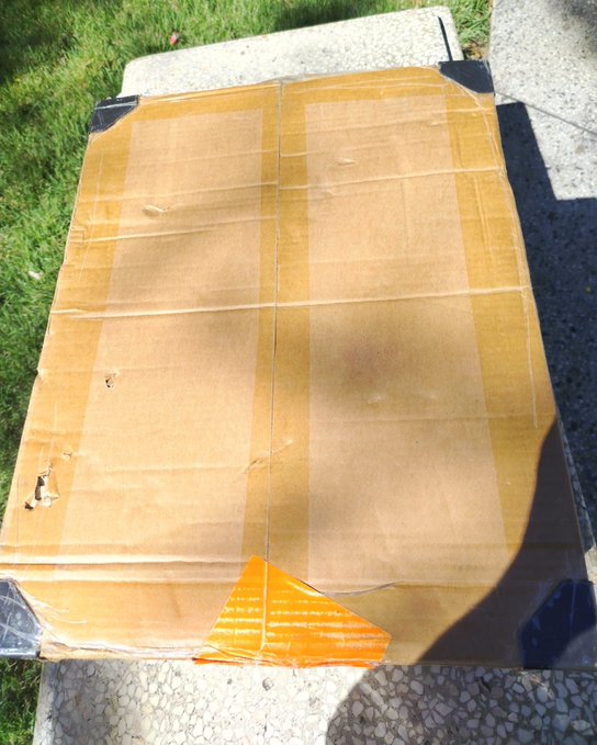 A cardboard-wrapped package sealed with brown packing tape and secured with black and orange tape, sitting on a concrete surface next to grass.