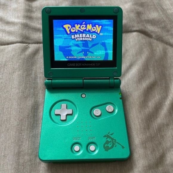 A green Game Boy Advance SP is shown displaying the title screen for 'Pokémon Emerald Version' on a fabric surface.