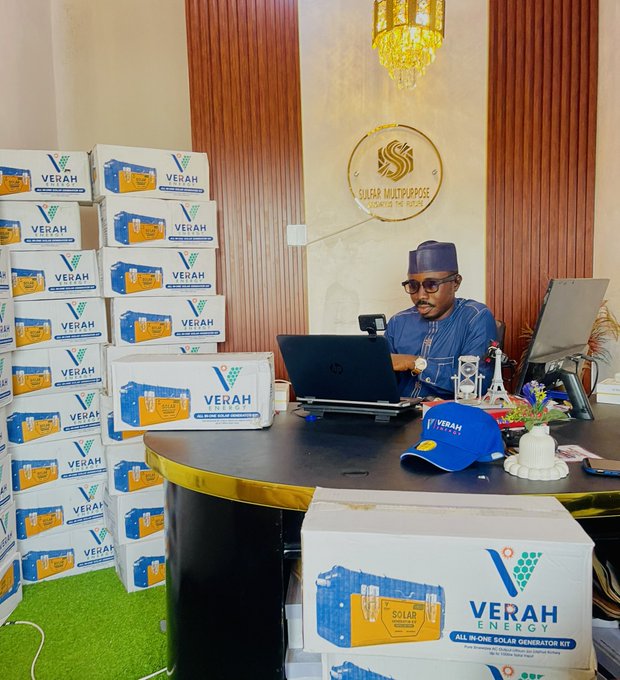 A man wearing a cap and glasses works on a laptop at a desk labeled "SUKAR MULTIPURPOSE," surrounded by stacks of boxes branded "VERAH ENERGY – All in One Solar Generator Kit," alongside a "SOLAR" cap and promotional materials on the desk.