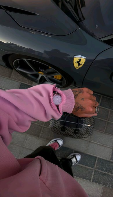 A person wearing a pink sweatshirt and sporting hand tattoos stands next to a sleek gray Ferrari, with the iconic prancing horse logo visible on the car's body. A black-and-white checkered duffel bag rests on the pavement near their red and white sneakers. The caption "gm ct don't lose your soul because of some sol" pairs with this image of luxury and style, encouraging followers to stay grounded and true to themselves despite external temptations or pressures.